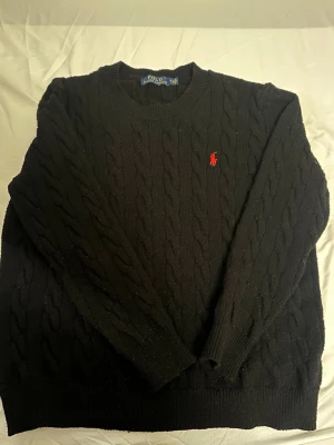 Svart kabelstickad tröja Polo Ralph Lauren - Kabelstickad svart tröja från Polo Ralph Lauren med klassisk rund halsringning och röd broderad logotyp på bröstet. Tröjan har långa ärmar och ribbade muddar. Perfekt för att hålla värmen och se stilren ut.