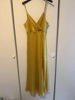 Gul långklänning med knappar - Snygg gul långklänning med smala axelband och omlottliv. Klänningen har dekorativa knappar längs ena sidan och en liten slits nedtill. Materialet har en lätt glansig finish som ger en lyxig känsla. Perfekt för sommarens alla tillfällen.