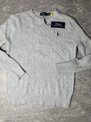Ljusgrå kabelstickad tröja Polo Ralph Lauren - Snygg ljusgrå kabelstickad tröja från Polo Ralph Lauren med rund halsringning och klassisk broderad logga på bröstet. Tröjan har ribbade muddar vid ärmslut och nederkant. Perfekt för dig som gillar stilrena och tidlösa plagg.