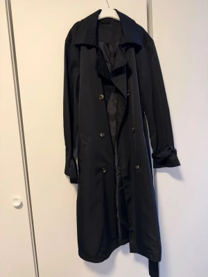 Svart klassisk trenchcoat - Stilren svart trenchcoat med dubbelknäppning och bred krage. Jackan har långa ärmar, knytskärp i midjan och knappar framtill. Perfekt för en trendig och tidlös look. Materialet ser ut att vara lätt och något glansigt.