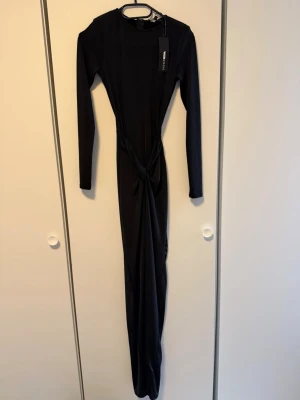 Svart långklänning  - Elegant svart långklänning med långärmad design och snygg knytning i midjan. Klänningen har en slank och figurnära passform som framhäver siluetten. Perfekt för dig som vill ha en stilren look med en twist.