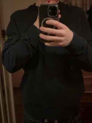 Svart sweatshirt från Stone Island - Säljer en svart sweatshirt från Stone Island med klassisk logotyp-patch på vänster ärm. Tröjan har rund halsringning, ribbade muddar och är tillverkad i mjukt material. Perfekt för en stilren och avslappnad look. Riktig bra skick, köpt från second place i Malmö 