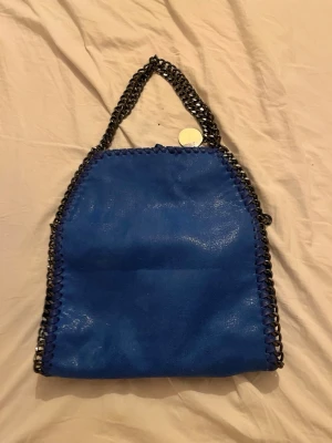 Blå Stella McCartney Falabella väska - Cool blå handväska från amazon. Väskan har en kedjerem i mörk metall och är gjord i veganskt läder med dekorativa sömmar längs kanterna. Kan bäras både i handen och över axeln tack vare den långa kedjan. Helt oanvänd.