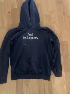 Mörkblå hoodie från Peak Performance - Snygg mörkblå hoodie från Peak Performance med vit logotyp på bröstet och klassisk känguruficka. Hoodien har ribbade muddar och är tillverkad i mjukt material som är perfekt för en avslappnad look.
