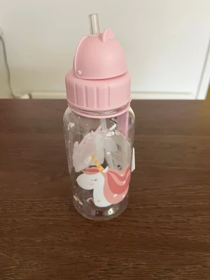 Rosa vattenflaska med enhörning - Söt vattenflaska med rosa lock och sugrör, dekorerad med en enhörning och små stjärnor. Flaskan är transparent och har ett lock som liknar ett djurhuvud. Perfekt för att ta med till skolan eller träningen.