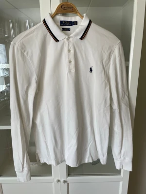 Vit långärmad piké från Ralph Lauren - Vit långärmad pikétröja från Polo Ralph Lauren med klassisk krage och knappar framtill. Slim fit-modell i bomull med stretch. Använd ett fåtal gånger. Ny pris: 849kr, mitt pris: 449kr 