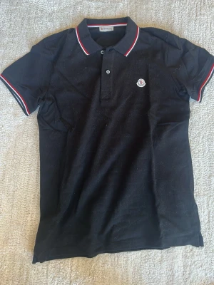 Svart pikétröja från Moncler - Svart pikétröja från Moncler med klassisk krage och knäppning framtill. Tröjan har röda och vita ränder på krage och ärmslut samt Moncler-logga på bröstet. Tillverkad i bomull och har korta ärmar. Perfekt för en stilren och sportig look.