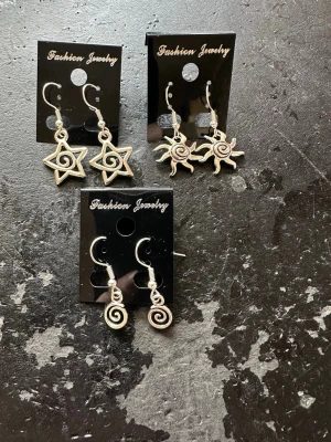 Set med tre par silvriga örhängen - Tre par coola silvriga örhängen i olika former: stjärnor med spiral, solar med spiral och spiraler. Alla har krokar i metall och en bohemisk vibe. Perfekta för dig som gillar unika accessoarer och vill sticka ut lite extra.
