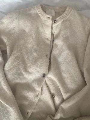 Beige kofta från H&M - Mysig beige kofta från H&M med rund halsringning och knappar framtill. Koftan är stickad i ett mjukt material och har långa ärmar. Perfekt att slänga på sig när det är lite kyligt ute.