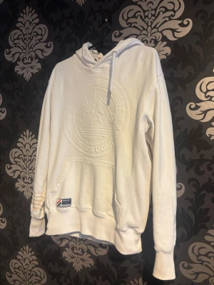 Vit hoodie från superdry - Säljer en vit hoodie från Napapijri med broderad logga på bröstet och klassisk känguruficka. Hoodien har huva med dragsko och ribbade muddar. Materialet är mjukt och bekvämt, perfekt för chill dagar. Patch med Napapijri-flagga på fickan.