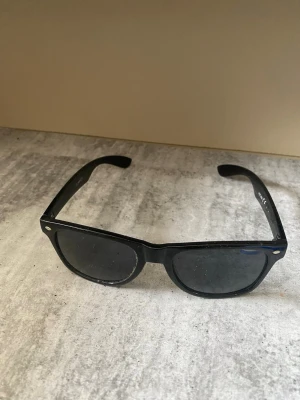 Svarta klassiska solglasögon - Säljer ett par svarta solglasögon med tidlös och klassisk form. Bågarna är i plast och har en enkel design med små silvriga detaljer vid hörnen. Perfekta för soliga dagar och ger en cool look.