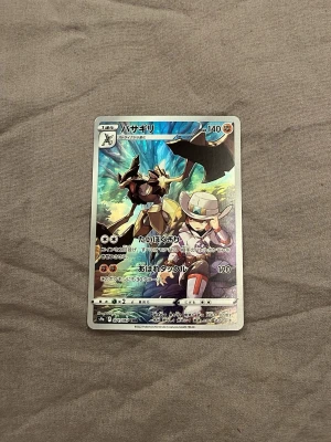 Japanskt Pokémonkort med holografisk effekt - Säljer ett japanskt Pokémonkort med holografisk yta och färgstark illustration. Kortet har motiv av en Pokémon och en tränare i en actionfylld scen. Texten är på japanska och kortet har detaljer i silver, blått och brunt.