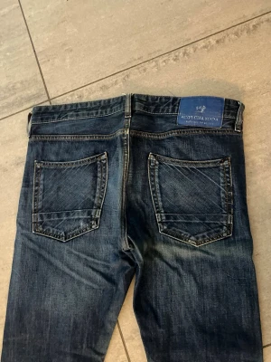 Mörkblå jeans från Scotch & Soda - Snygga mörkblå jeans från Scotch & Soda med klassisk femficksmodell och slitna detaljer. Jeansen har raka ben och en blå patch med logga bak i midjan. Tillverkade i denim med kontrastsömmar och cool tvätt. Storlek Ca: w:33 L:32, fråga om mått så skickar jag. 
