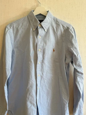 Ljusblå skjorta från Polo Ralph Lauren - Klassisk ljusblå skjorta från Polo Ralph Lauren i modellen Classic Fit. Skjortan har button-down krage, långa ärmar och den ikoniska broderade loggan på bröstet. Tillverkad i bomull för en skön och stilren look.