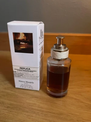 Maison Margiela  By the Fireplace - Maison Margiela  By the Fireplace är en parfym i en genomskinlig glasflaska med metallfärgad spray och vit textil runt halsen. Doften är varm och rökig, inspirerad av en brasa. Flaskan rymmer 30 ml och kommer i en vit kartong med bild på en eld. 20 ml kvar i flaskan. Köpt på kicks. Nypris 800kr.