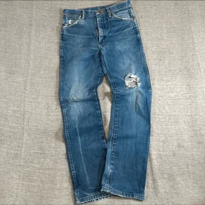 Blå raka jeans med slitningar - Säljer ett par klassiska blå jeans med raka ben och slitningar, inklusive ett större hål på ena låret. Jeansen har fem fickor, bälteshällor och en vintagekänsla. Perfekta för dig som gillar en avslappnad och trendig stil.