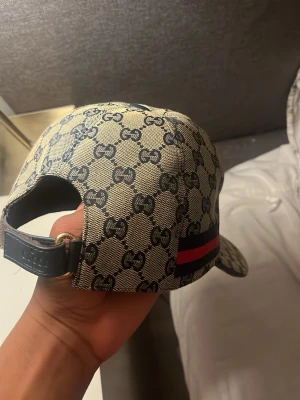  keps med GG-mönster - Snygg keps frånmed klassiskt GG-monogram i beige och mörkblått. Kepsen har justerbar rem baktill och en ikonisk röd och marinblå rand på sidan. Tillverkad i canvasmaterial som ger en exklusiv känsla.