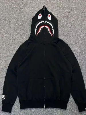 Svart hoodie med hajmotiv på huvan - Cool svart hoodie från BAPE med dragkedja och stor huva som har ett hajansikte med tänder och ögon tryckt på. Hoodien har ribbade muddar, fickor fram och en vit detalj på ärmen. Perfekt för streetwear-stilen.