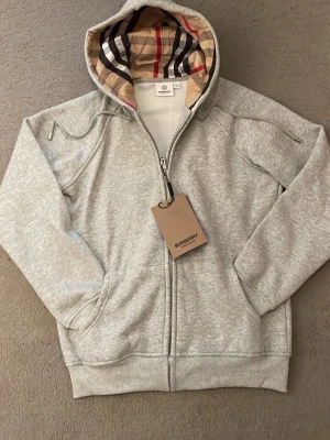 Grå hoodie med dragkedja från Burberry - Snygg grå hoodie från Burberry med dragkedja och klassiskt rutigt foder i huvan. Mjuk bomullsmix, ribbade muddar och fickor framtill. Perfekt för en stilren och avslappnad look.