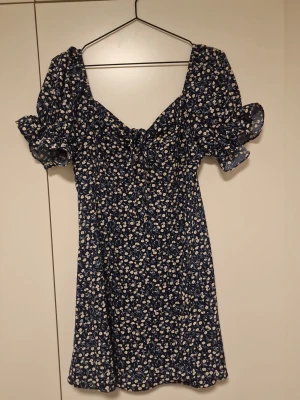 Blommig blå klänning med puffärm - Supersöt blå klänning med små vita blommor och puffiga korta ärmar. Klänningen har en lätt v-ringning och en figurnära passform upptill. Perfekt för varma dagar och har en romantisk vibe.