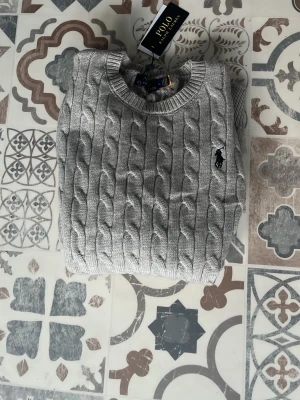 Grå kabelstickad tröja Polo Ralph Lauren - Snygg grå kabelstickad tröja från Polo Ralph Lauren med rund halsringning och broderad logga på bröstet. Tröjan har långa ärmar och ribbade muddar vid ärmslut och nederkant. Perfekt för lager-på-lager och klassisk stil.