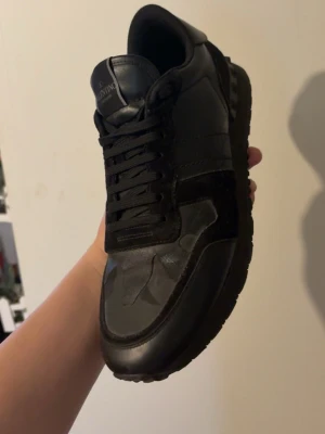 Svarta Valentino Garavani sneakers - Snygga svarta sneakers från Valentino Garavani med diskret kamouflagemönster och detaljer i mocka och läder. Klassisk låg modell med svarta skosnören och logga på plösen. Perfekta för dig som gillar stilrena och exklusiva sneakers.