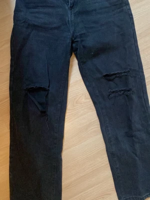 Svarta ripped jeans med hål på knäna - Snygga svarta jeans med trendiga slitningar och hål på båda knäna. Modellen har raka ben och klassisk femficksdesign. Perfekta för dig som gillar en edgy och avslappnad stil.