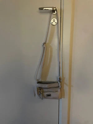 Beige axelväska från Feel - Snygg beige axelväska från Feel med justerbar rem och två dragkedjefack framtill. Väskan är i ett mjukt konstläder och har silverfärgade detaljer. Perfekt storlek för det viktigaste och enkel att matcha till olika outfits.