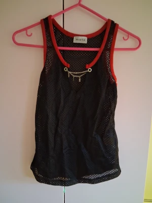 Svart mesh linnetopp med kedjedetalj - Cool svart mesh linnetopp från Michi med röda kanter och en snygg kedjedetalj framtill. Perfekt för dig som vill sticka ut med sportig och edgy stil. Luftigt material och ärmlös design gör den superbekväm.