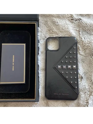 Svart mobilskal med nitar Ideal of Sweden - Snyggt svart mobilskal från Ideal of Sweden med silverfärgade nitar i olika former på baksidan. Skalet har en stilren design med en diagonal detalj och är gjort i konstläder. Perfekt för dig som vill ha ett trendigt och unikt skydd till din mobil.