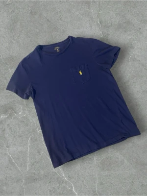 Blå t-shirt från Polo Ralph Lauren | S - Snygg marinblå t-shirt från Polo Ralph Lauren med klassisk gul logga broderad på bröstfickan. T-shirten har rund halsringning och korta ärmar. Tillverkad i mjuk bomull för en bekväm känsla. Perfekt till jeans eller shorts.