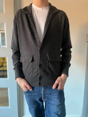 Massimo dutti tröja  - Mysig mörkgrå kofta med knappar framtill och två stora fickor med lock. Koftan har ribbade muddar och en klassisk krage. Perfekt att slänga över en t-shirt för en avslappnad look. Materialet ser ut att vara mjukt och stickat. Storlek M.