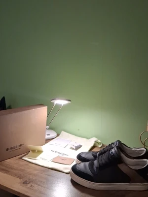 Burberry svarta och bruna sneakers - Stilrena sneakers från Burberry i svart och brunt läder med mockadetaljer och klassisk logga på plösen. Skorna har vita sulor och lågt skaft, samt paneler i textil på sidorna. Perfekta för dig som gillar exklusiva och trendiga märken. Är använt för nån halv-1 månad men har INGA DEFEKTER.