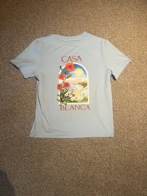 Casa blanca - Snygg ljusblå t-shirt från Casa Blanca med stort färgglatt tryck på ryggen som visar rosor och ett somrigt landskap. T-shirten har korta ärmar. Perfekt för dig som gillar unika prints och vill sticka ut lite extra.