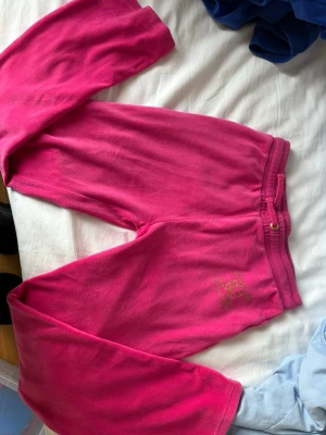 Juicy Couture rosa mjukisbyxor - Säljer ett par klassiska rosa mjukisbyxor från Juicy Couture med broderad logga i guld på ena benet. Byxorna har resår i midjan och är gjorda i mjuk velour som är superbekväm. Perfekta för en chill och trendig look.