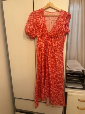 Röd långklänning med vita prickar - Röd långklänning med vita prickar och v-ringad hals. Klänningen har puffiga korta ärmar, knappar framtill och ett skärp i midjan för snygg passform. Perfekt för sommarens alla tillfällen.
