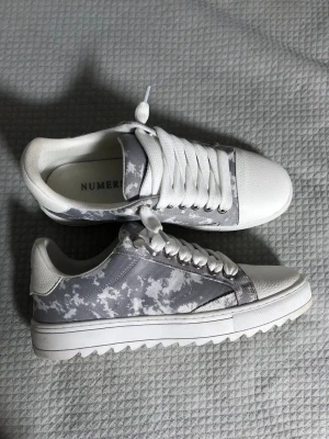 Gråvita sneakers från numeris - Snygga sneakers från numeris i vitt och grått med camo-mönster på sidan. Ovandelen är i textil och syntet, med vita snören och detaljer. Sulan är vit och har ett räfflat mönster för extra grepp. Perfekta för en clean och modern stil.