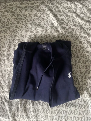Mörkblå hoodie från Polo Ralph Lauren - Snygg mörkblå hoodie från Polo Ralph Lauren med dragkedja, huva med dragsko och två fickor framtill. Ikoniska vita loggan broderad på bröstet. Perfekt för en chill och stilren look.