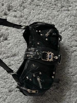 Svart väska med nitar och detaljer - Cool svart handväska i glansigt syntetisk material med flera dragkedjor, silverfärgade nitar och en unik spänne med ögonformad dekoration. Väskan har justerbart axelband och edgy design som sticker ut.