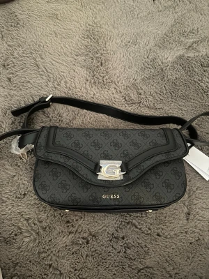 Svart axelväska från Guess - Snygg svart axelväska från Guess med justerbar rem och silverfärgad logga framtill. Väskan har mönstrat konstläder med Guess-loggor och flera fack inuti för smart organisering. Perfekt accessoar för att lyfta din outfit.