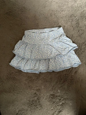 Blommig volangkjol från Lindex - Supersöt ljusblå kortkjol från Lindex med vitt blommönster och dubbla volanger. Kjolen har en bekväm resår i midjan och är perfekt för vår och sommar. Tillverkad i mjuk bomull som känns skön mot huden.❣️❣️