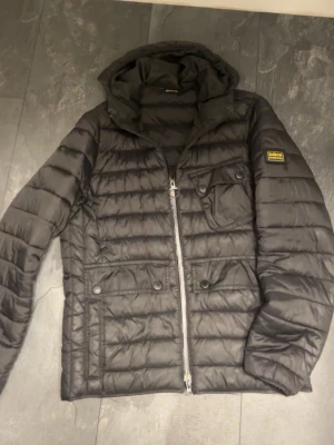 Svart quiltad pufferjacka Barbour SKRIV INNAN KÖP! - Snygg svart pufferjacka från Barbour med quiltad design, huva och flera fickor med knappstängning. Jackan har dragkedja framtill och en patch med Barbour-logga på ärmen. Perfekt för kyliga dagar och enkel att matcha.