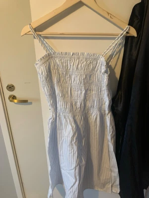 Ljusblå- och vitrandig sommarklänning - Säljer en söt kort klänning med smala axelband och smock upptill. Klänningen är ljusblå- och vitrandig, har en avslappnad passform och är perfekt för varma dagar. Tillverkad i mjukt bomullstyg som känns skönt mot huden.