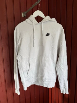 Vit Nike hoodie med logga - Snygg vit hoodie från Nike med svart broderad logga på bröstet. Klassisk modell med huva och dragsnören samt stor känguruficka framtill. Perfekt för en avslappnad streetstyle-look. Tillverkad i mjukt material för extra komfort.
