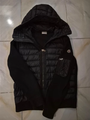 Svart pufferjacka från Moncler - Snygg svart pufferjacka från Moncler med stickade ärmar och huva. Jackan har dragkedja framtill, en ficka med dragkedja på ärmen och Moncler-logga på vänster ärm. Materialmix med quiltad front och stickade detaljer ger en cool look.