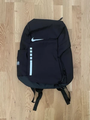 Svart Nike Elite ryggsäck - Snygg svart Nike Elite ryggsäck med vit logga och reflekterande detaljer. Ryggsäcken har flera fack, vadderade axelband med ELITE-tryck och sidoficka. Tillverkad i slitstarkt syntetmaterial, perfekt för skola eller träning. Helt ny med prislapp.