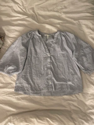 Randig blus med knappar från H&M - Blå- och vitrandig blus från H&M med knappar framtill och trekvartsärmar. Blusen har en avslappnad passform och är tillverkad i ett lätt bomullstyg. Perfekt för en fräsch och stilren look.