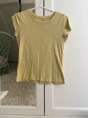 Gul t-shirt från Polo Ralph Lauren - Snygg gul t-shirt från Polo Ralph Lauren med klassisk rund hals och liten broderad logga på bröstet. Tillverkad i mjuk bomull och har korta ärmar. Perfekt för en avslappnad och stilren look.