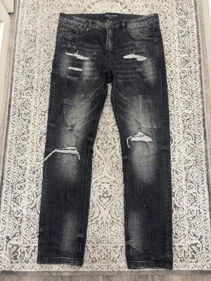 Svarta slitna jeans från Supply and Demand - Säljer ett par svarta jeans från Supply & Demand med slitningar och trasiga detaljer på benen. Jeansen har en smal passform och klassisk femficksdesign. Snyggt tvättade partier och coola slitningar ger en trendig look. Perfekta för dig som gillar streetstyle.