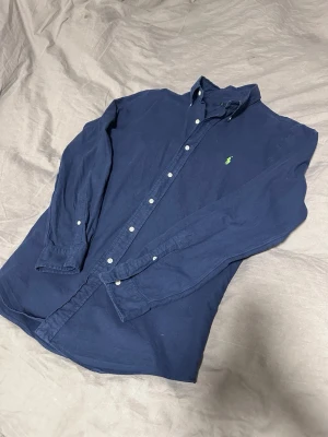 Mörkblå skjorta från Ralph Lauren - Klassisk mörkblå skjorta från Ralph Lauren med button-down krage och vita knappar. Skjortan har lång ärm och den ikoniska gröna broderade loggan på bröstet. Tillverkad i mjukt bomullstyg som känns skönt mot huden.
